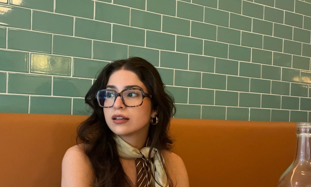 woman sitting at a booth wearing oversized glasses and a scarf tied around her neck